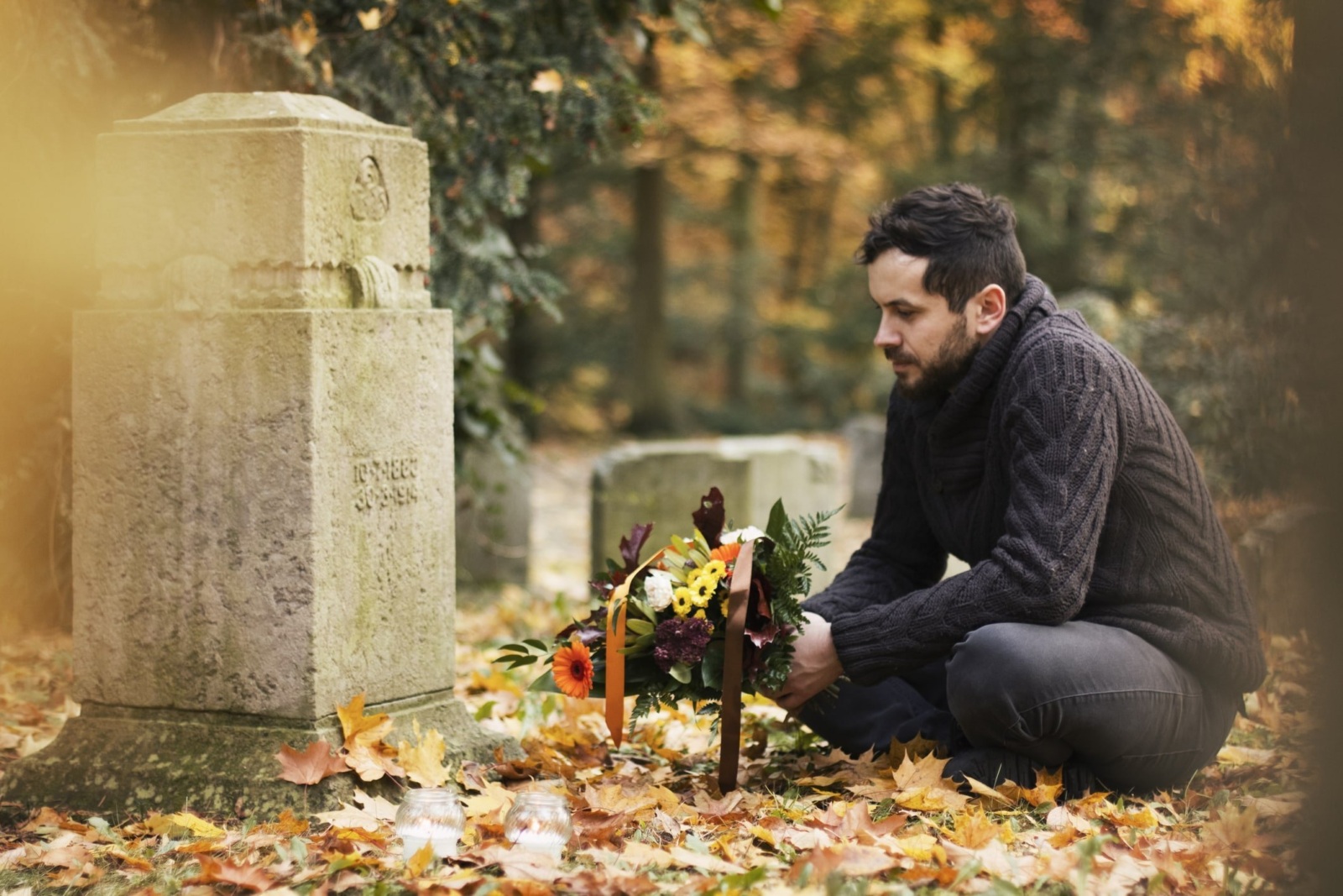 A man holding a bouquet of flowers sits beside a gravestone surrounded by autumn leaves.