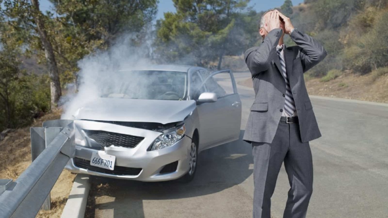 A man in a suit stands in distress next to a damaged car with smoke coming from the hood after a crash into a guardrail.