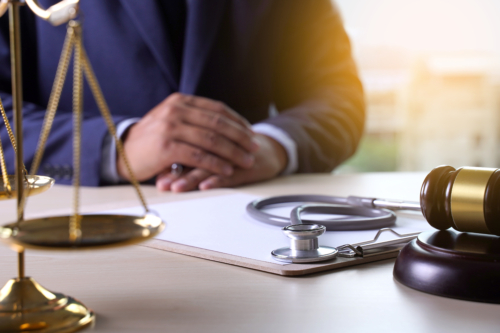 A lawyer sits at their desk facing a clipboard, stethoscope, gavel and sound block, and the scales of justice.