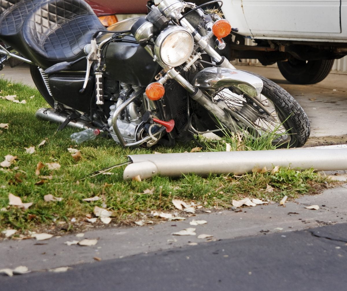 A motorcycle lies in the grass after a collision with another vehicle which is seen in the background.