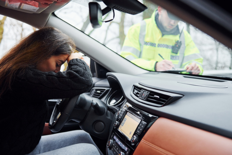 Female driver with head and hands on steering wheel while officer writes her a ticket.