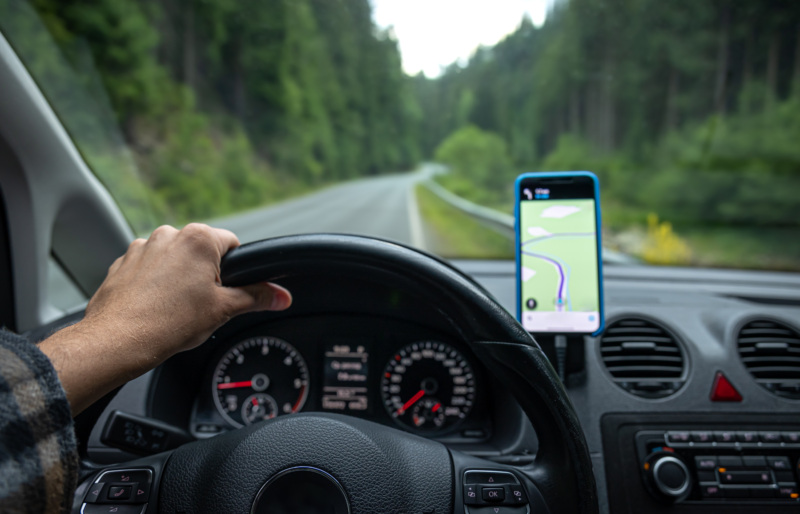 Driver view of steering wheel, dashboard, road, and phone displaying navigation.