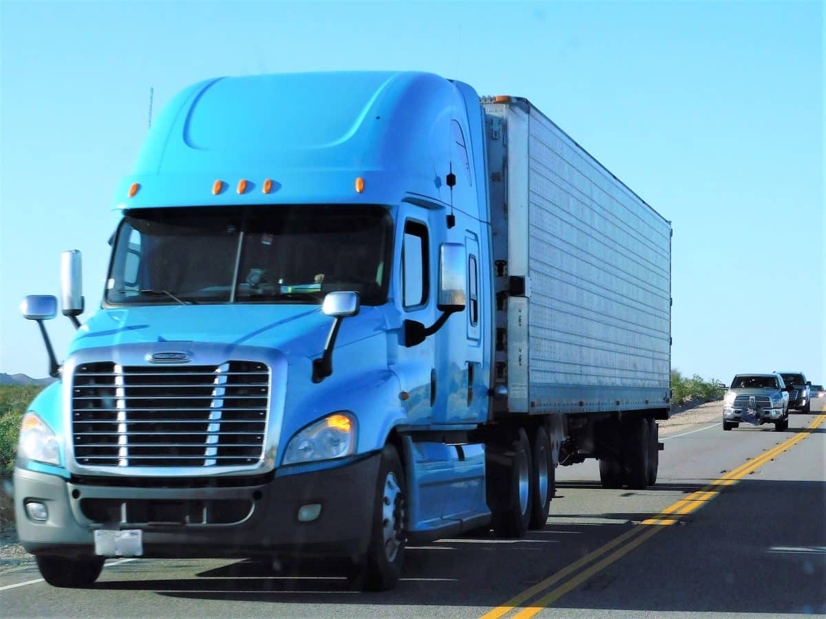 A blue semi-truck drives on the highway followed by 3 passenger vehicles.