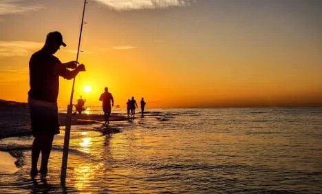 A group of people fishing and walking along the beach at sunset.