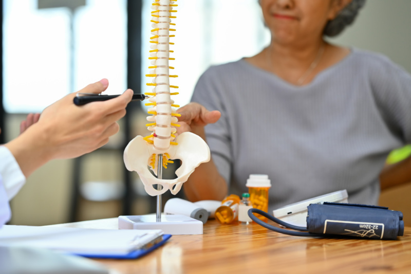 An orthopedist pointing at a spine model while talking to a patient at a desk.