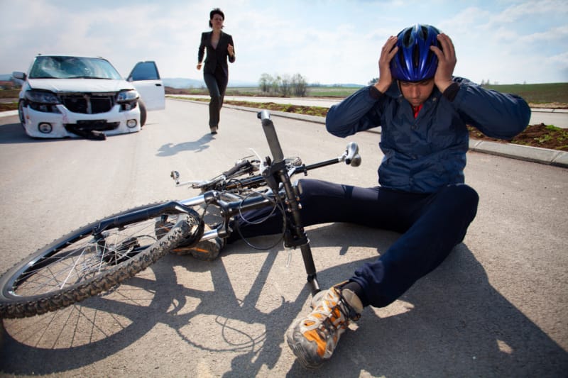 A woman runs from her car to an injured bicyclist who is sitting on the ground next to his bike, holding his head.