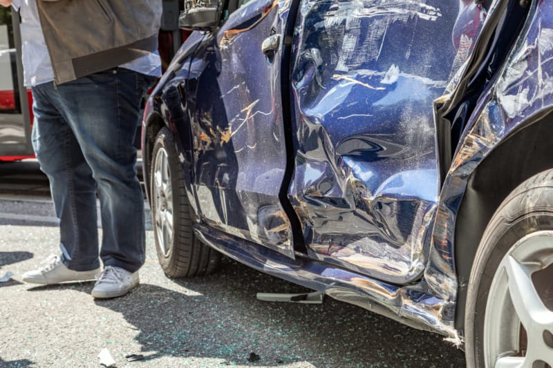 View of a man from the midsection down, standing next to a blue sedan with dented left passenger and driver doors.