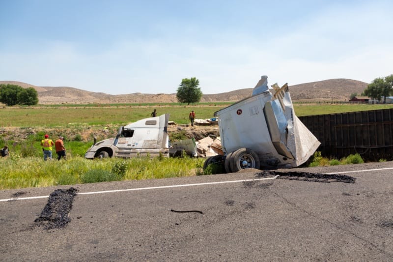 Construction workers gather around a big rig on the side of the road that has suffered significant damage in an accident.