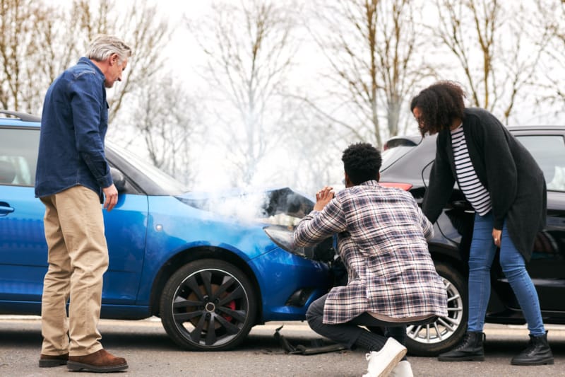 A rideshare driver, their passenger, and another driver examine the damage from a rear-end collision.