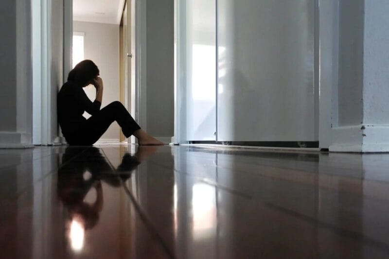 A woman sits on the floor of her home with her head in her hands.
