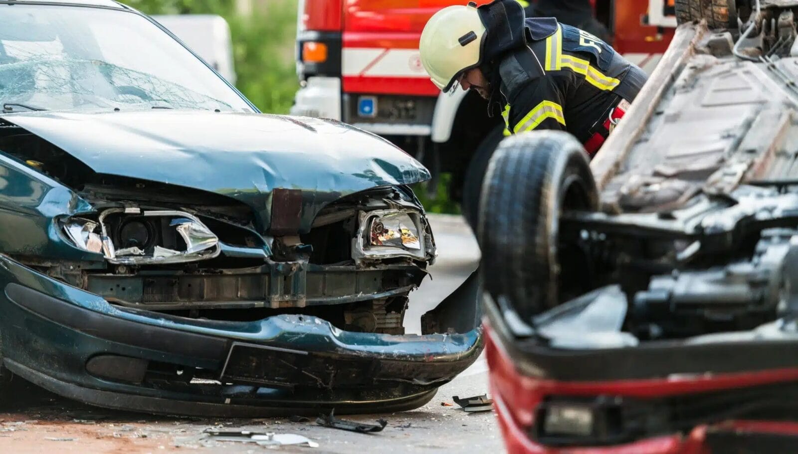A fireman examines the damage to the front of a black sedan. A red sedan lies overturned in the foreground.