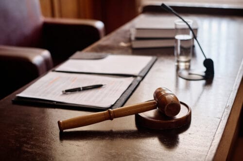 A wooden gavel sits on a sound block beside an open binder and legal documents on a judge’s desk.
