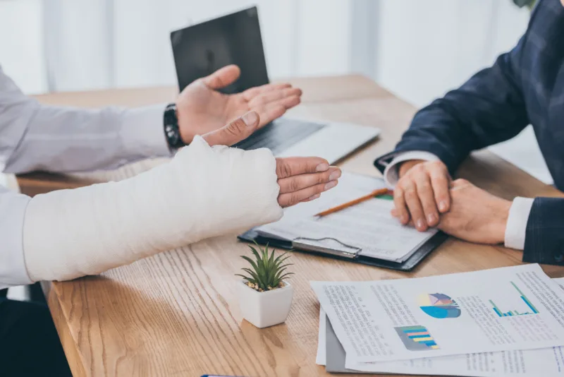 Cropped view of an individual with a broken arm sitting at a desk discussing their personal injury case with their lawyer